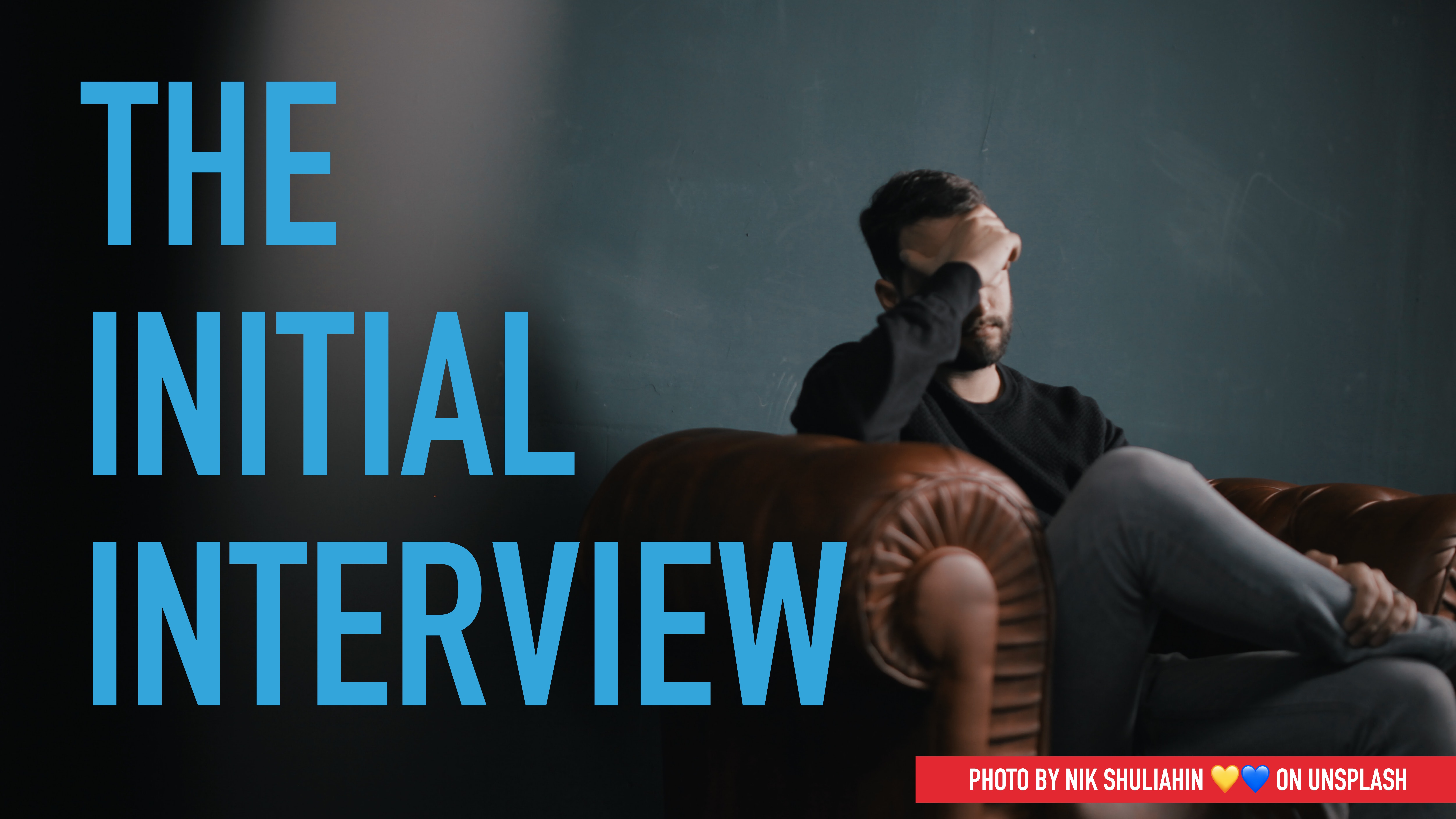 A person sits on a brown leather sofa, holding their forehead, set against a dimly lit room. Large blue text reads 'THE INITIAL INTERVIEW.' Photo credit: Nik Shuliahin on Unsplash.